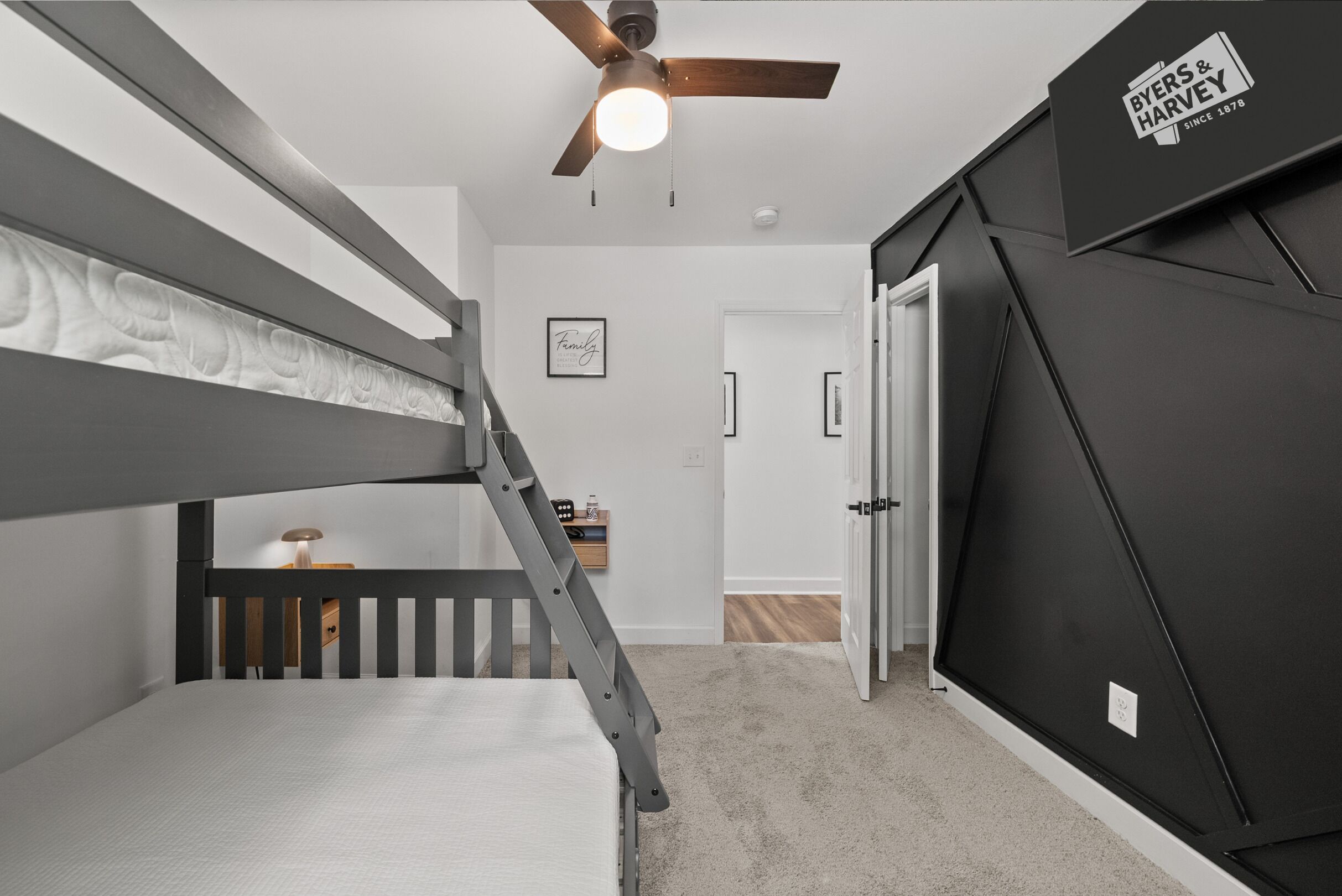 A room with a bunk bed, a ceiling fan, a framed family quote on the wall, and a door leading to another room.