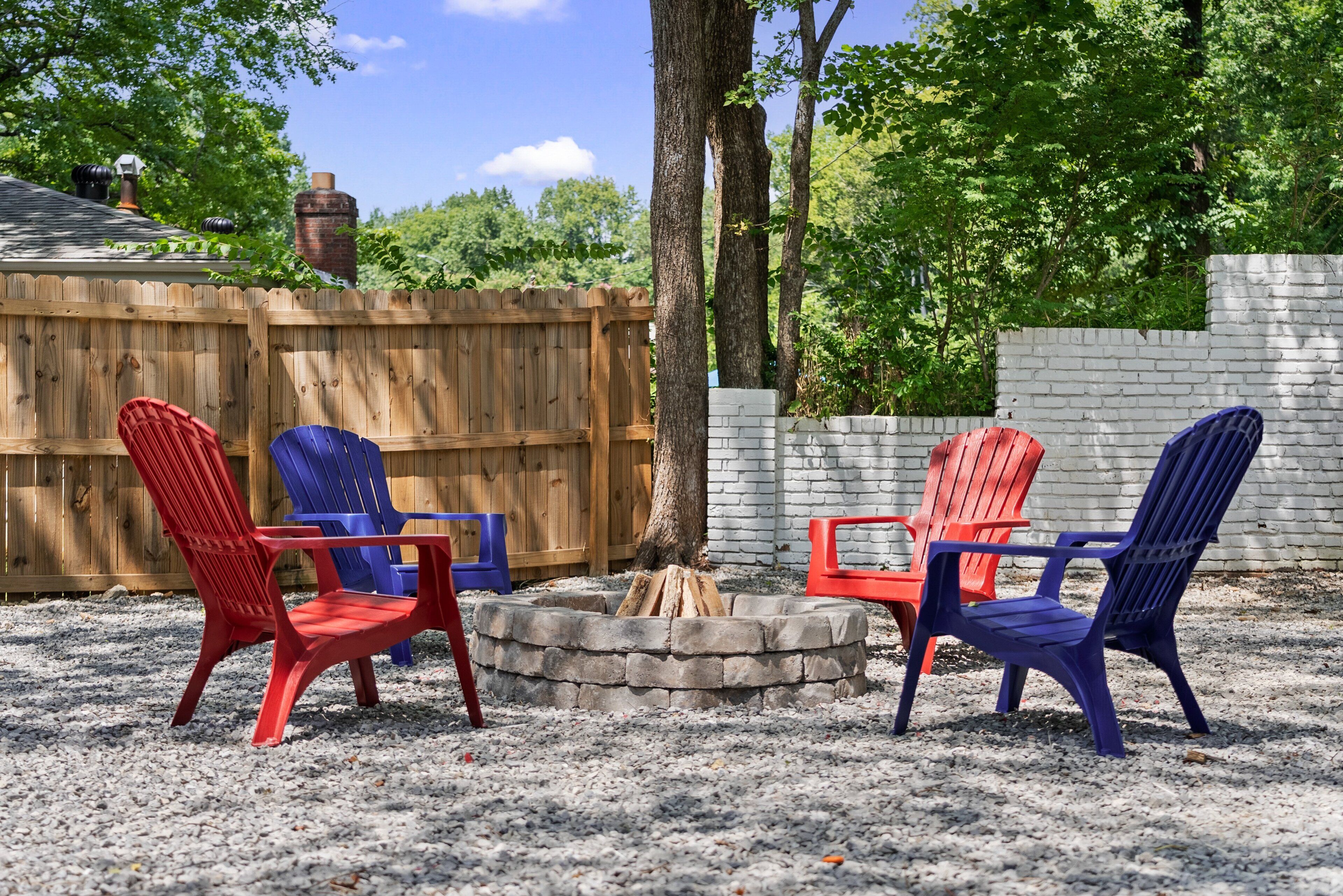 A backyard with a wooden fence, a brick wall, and a fire pit surrounded by red and blue Adirondack chairs.