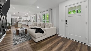A modern living room with a sofa, a coffee table, and a rug. There is a kitchen in the background with cabinets and a refrigerator. - Bertin's Place (Clarksville)