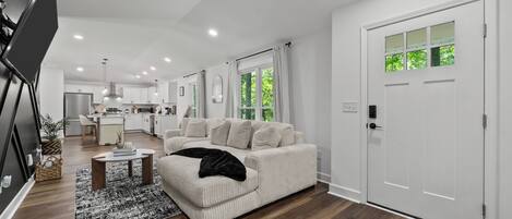 A modern living room with a sofa, a coffee table, and a rug. There is a kitchen in the background with cabinets and a refrigerator.