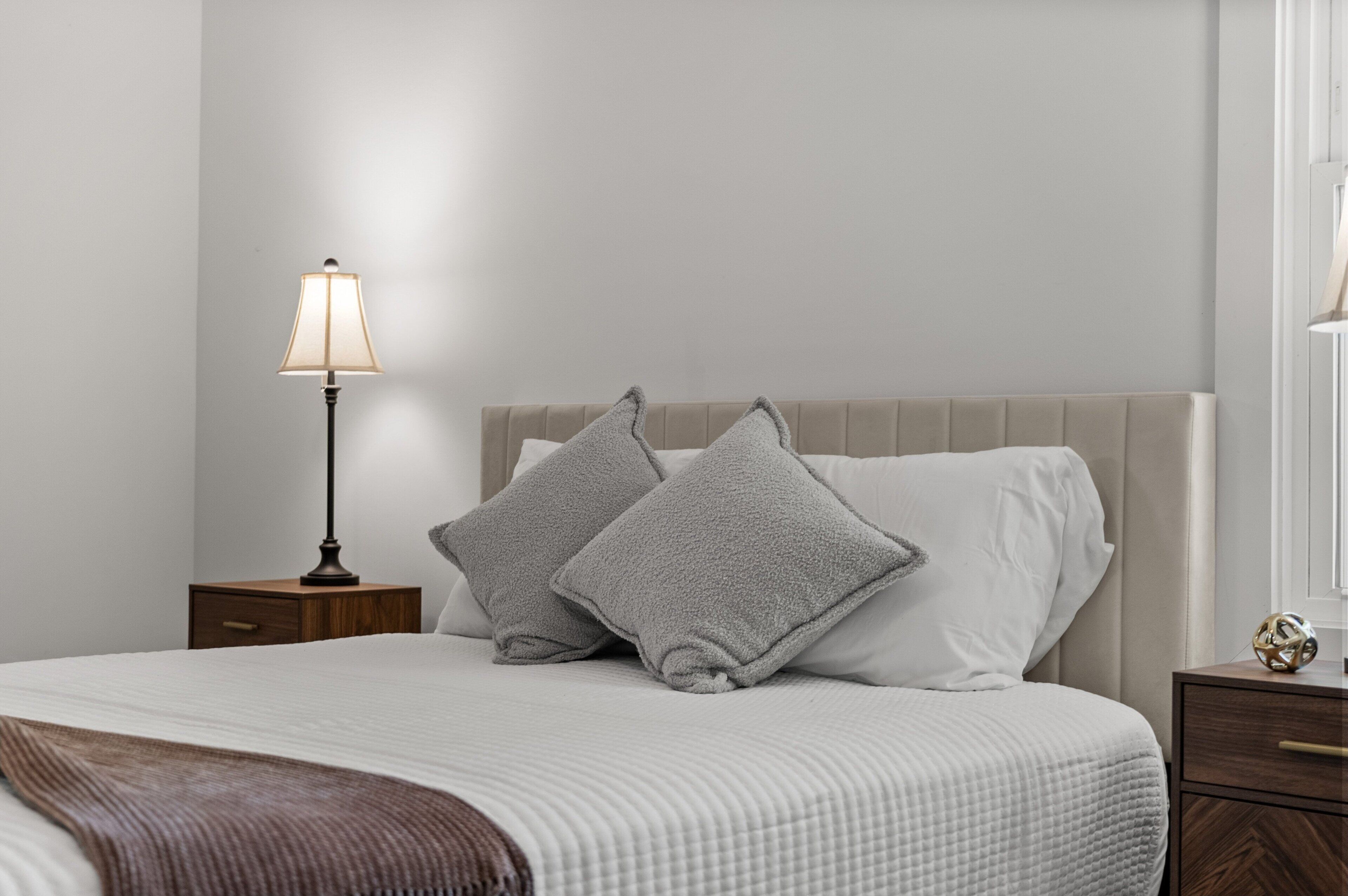 Un letto rifatto con un copriletto bianco e due cuscini grigi, una lampada da comodino e un comodino in legno con un cassetto.