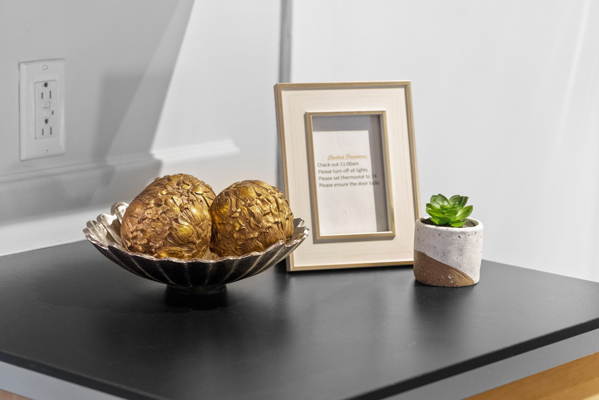 A black countertop with a decorative bowl containing golden objects, a framed sign with instructions, and a small potted plant.