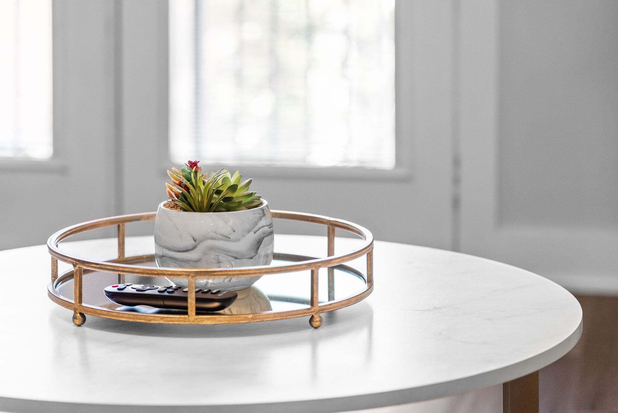 A round table with a white marble top, a white marble vase with a succulent plant, and a remote control on a gold-framed tray.