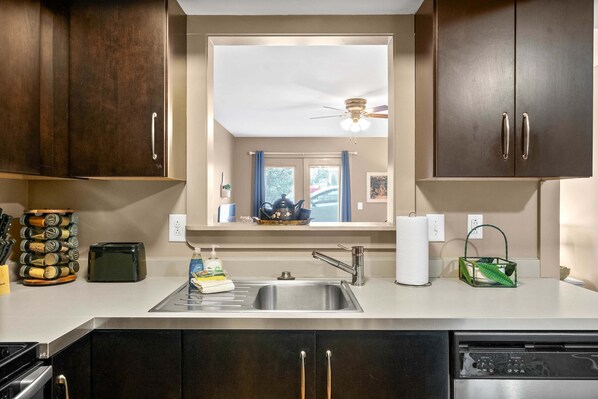 A kitchen with dark wooden cabinets, a stainless steel sink, a ceiling fan, and a window with blue curtains.