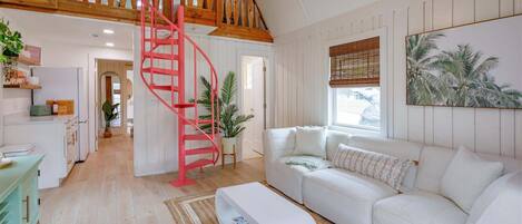 A spacious living area with a white sofa, a red spiral staircase, and a large window with a view of palm trees.