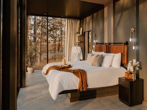 A modern bedroom with a large bed, a bedside table, a nightstand, and a window with a view of trees. - Whisper Mountain Glass Suite! Steam Shower-Couples Tubs-Bidet-Panoramic Views (Broken Bow)