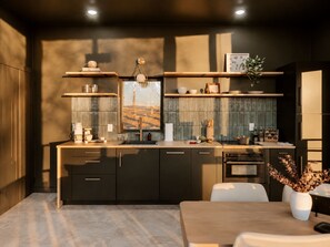 A modern kitchen with dark cabinetry, a central island, and a dining area with a table and chairs. - Whisper Mountain Glass Suite! Steam Shower-Couples Tubs-Bidet-Panoramic Views (Broken Bow)