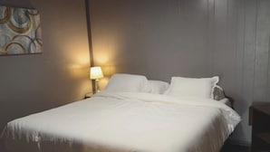 A neatly made bed with white linens in a room with wooden walls and a single bedside table with a lamp.