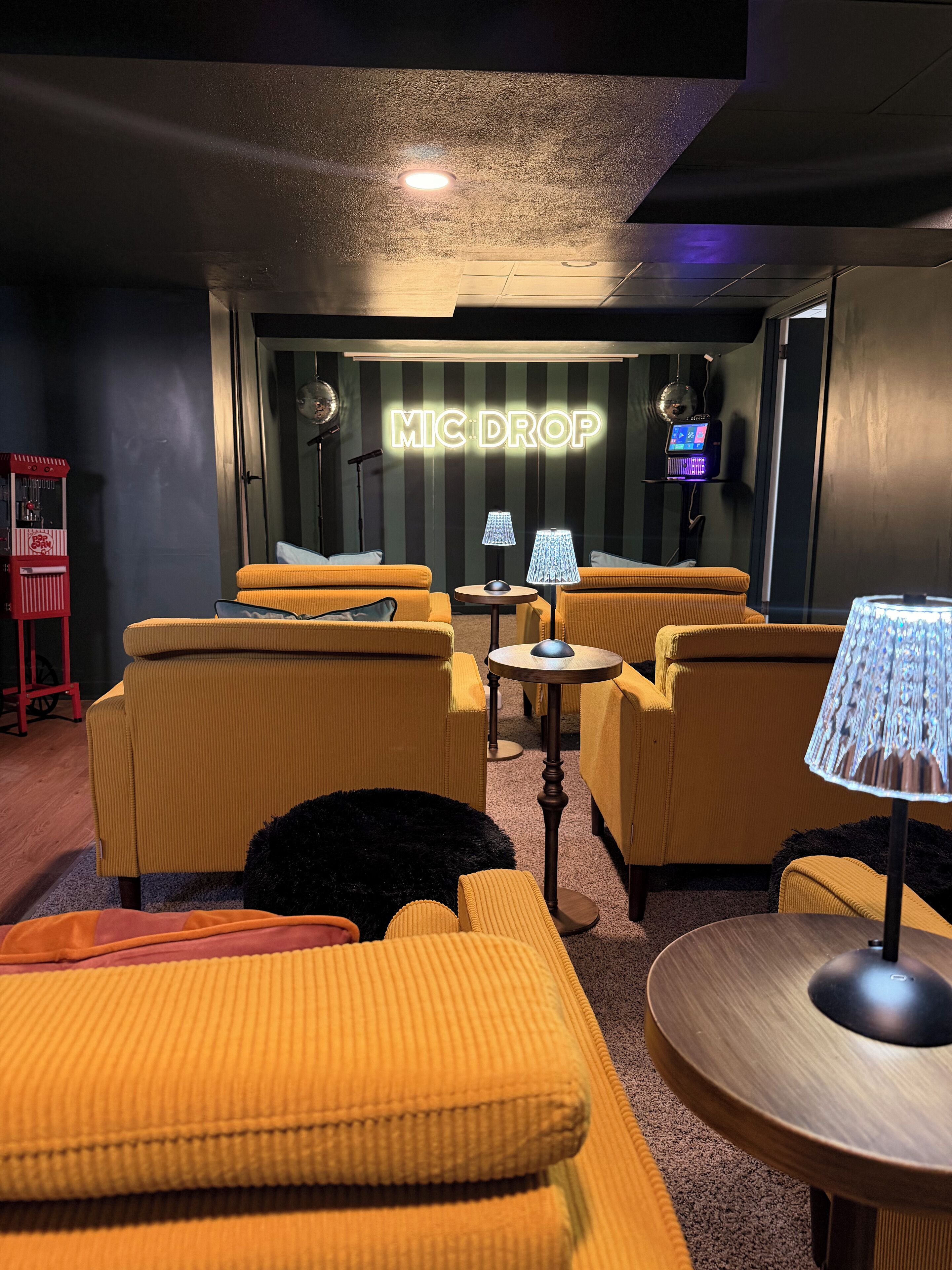 A cozy lounge area with yellow upholstered sofas, a small round table, and a lamp. A neon sign reads "MIC DROP" on the wall.