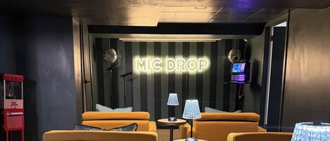 A cozy lounge area with yellow upholstered sofas, a small round table, and a lamp. A neon sign reads "MIC DROP" on the wall.