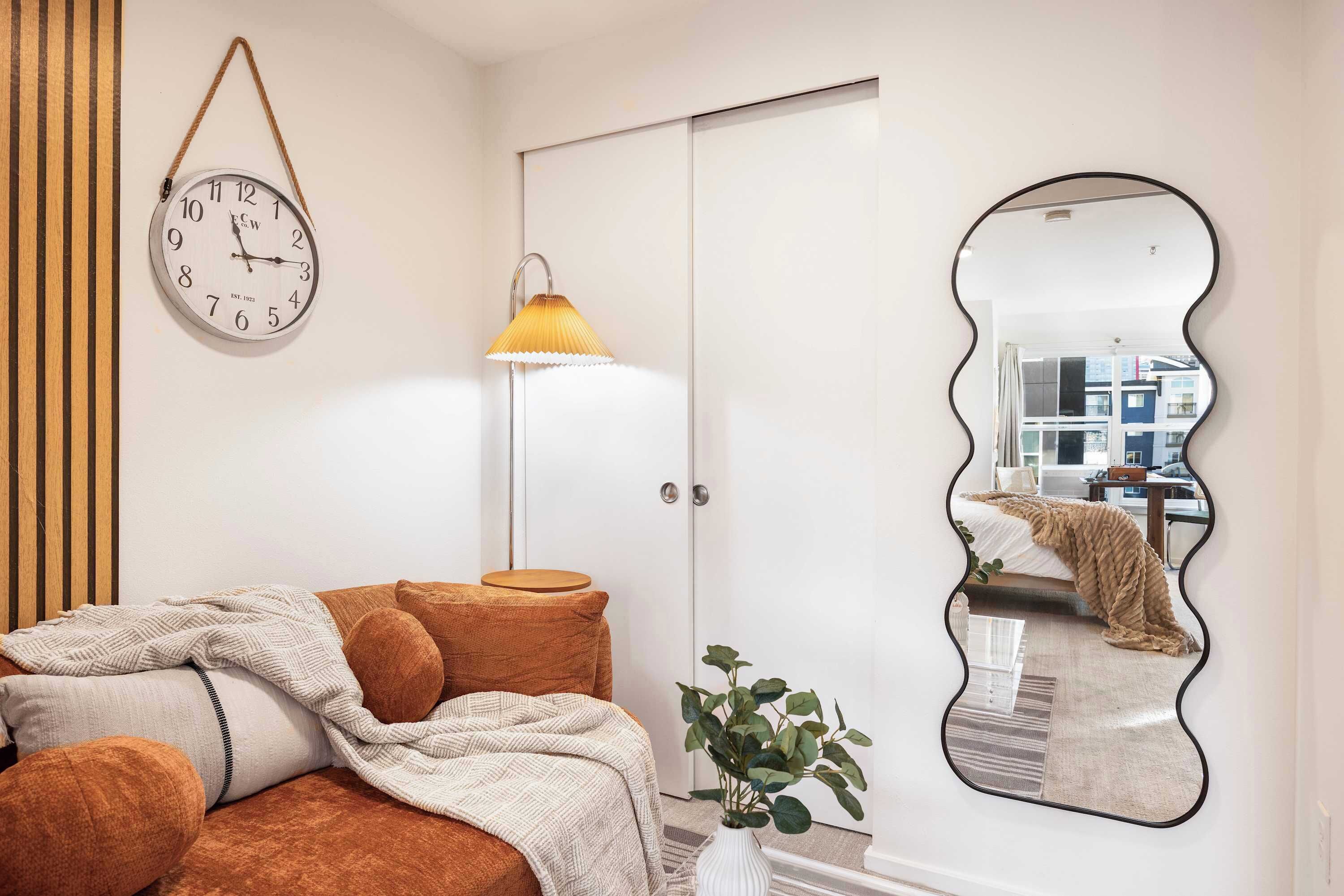 A bedroom with a bed, a bedside lamp, a wall clock, a mirror, and a plant.