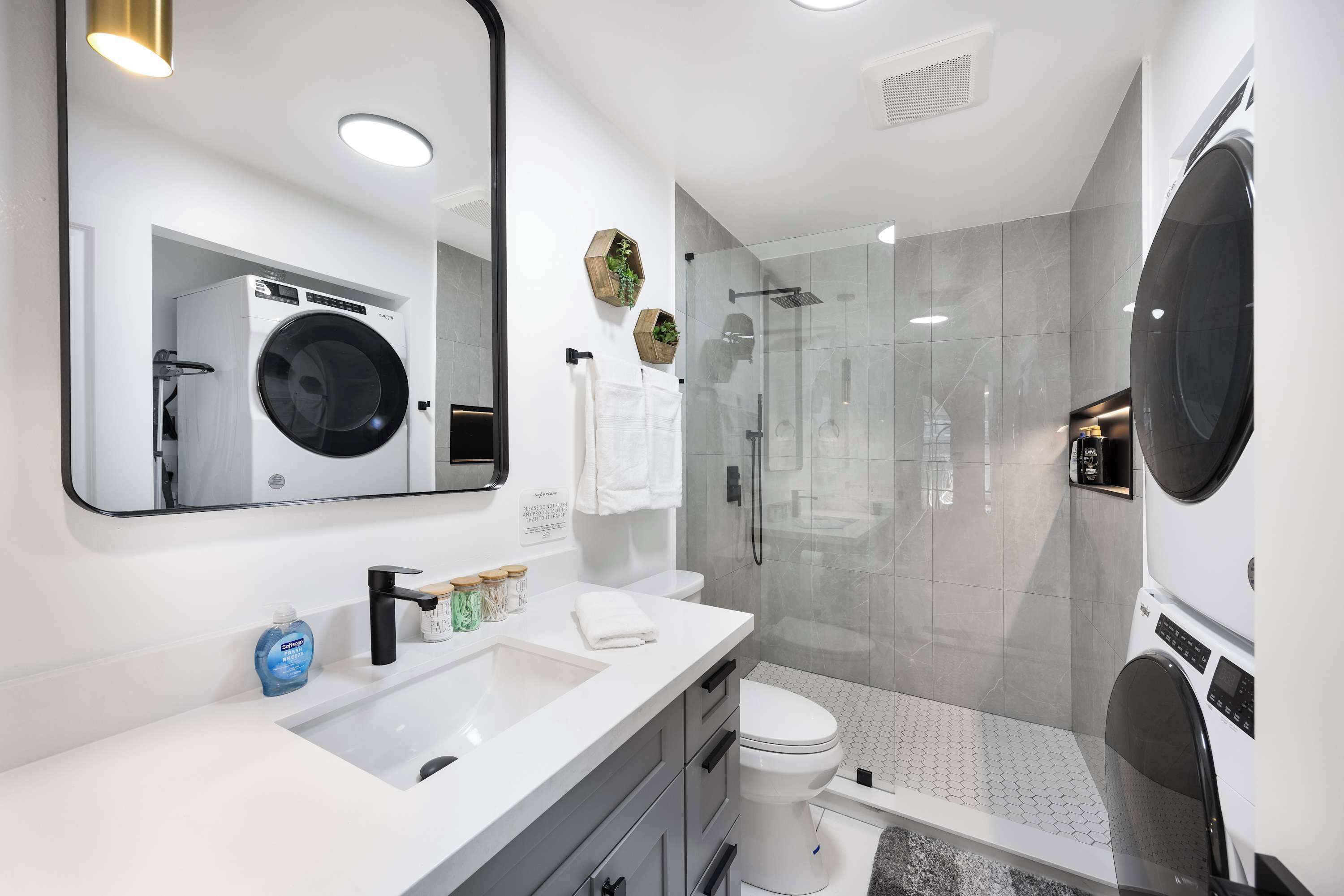 Un baño moderno con lavadora y secadora integradas, cabina de ducha de vidrio y tocador con lavamanos y espejo.