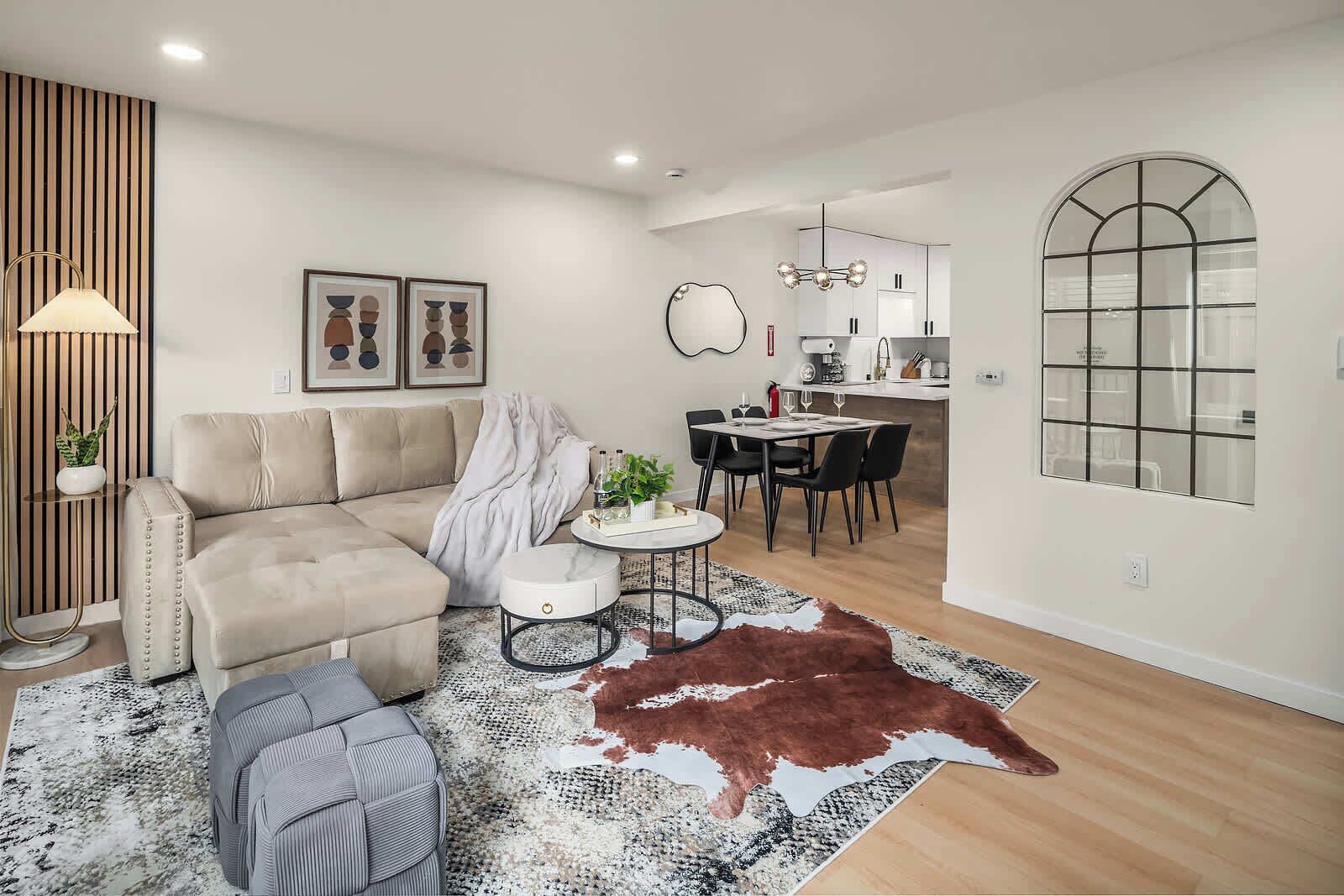 A modern living room with a sofa, a round coffee table, and a dining area with a table and chairs. There is a kitchen in the background with a window and a pendant light.