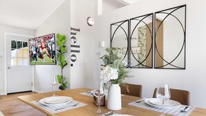 Een eetruimte met een houten tafel gedekt voor vier personen, witte borden en wijnglazen. Aan de muur hangt een groot sportposter in lijst en een geometrisch spiegel.