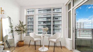 Un apartamento moderno con un ventanal grande, una mesa redonda, dos sillas blancas, una planta en maceta y un espejo grande.