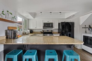 Una cocina moderna con armarios blancos, encimera de granito, microondas integrado, refrigerador de acero inoxidable y tres taburetes azules.