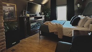 Et hyggeligt soveværelse i loftstil med skrå vægge, en ventilator i loftet, en seng med et beige tæppe, en sort lædersofa og et fjernsyn på et træstel.