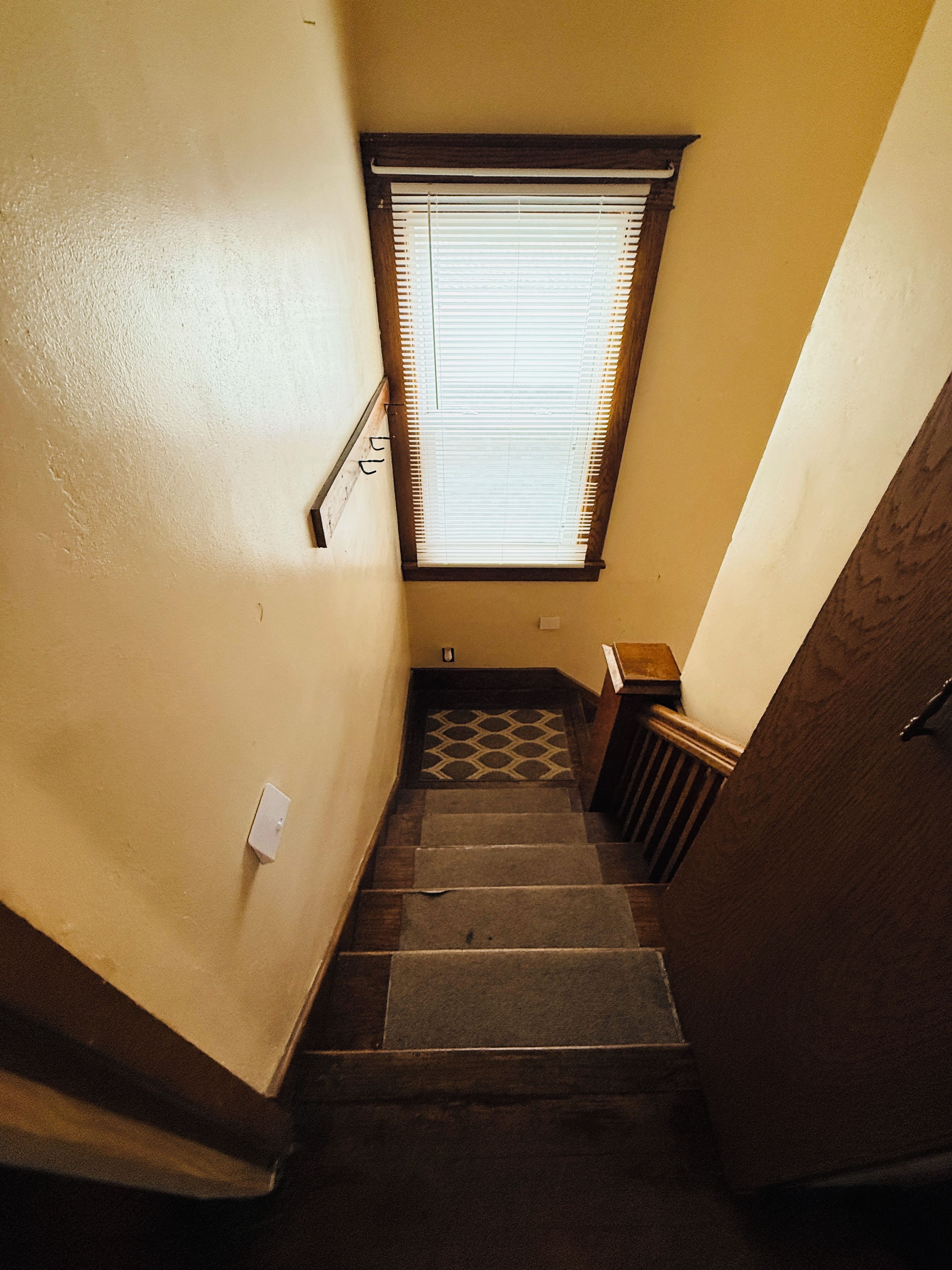 A narrow stairway with a wooden handrail, a window with blinds, and a patterned floor.