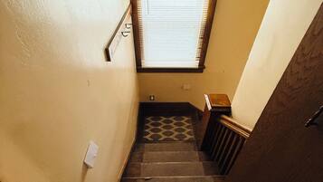 A narrow stairway with a wooden handrail, a window with blinds, and a patterned floor.