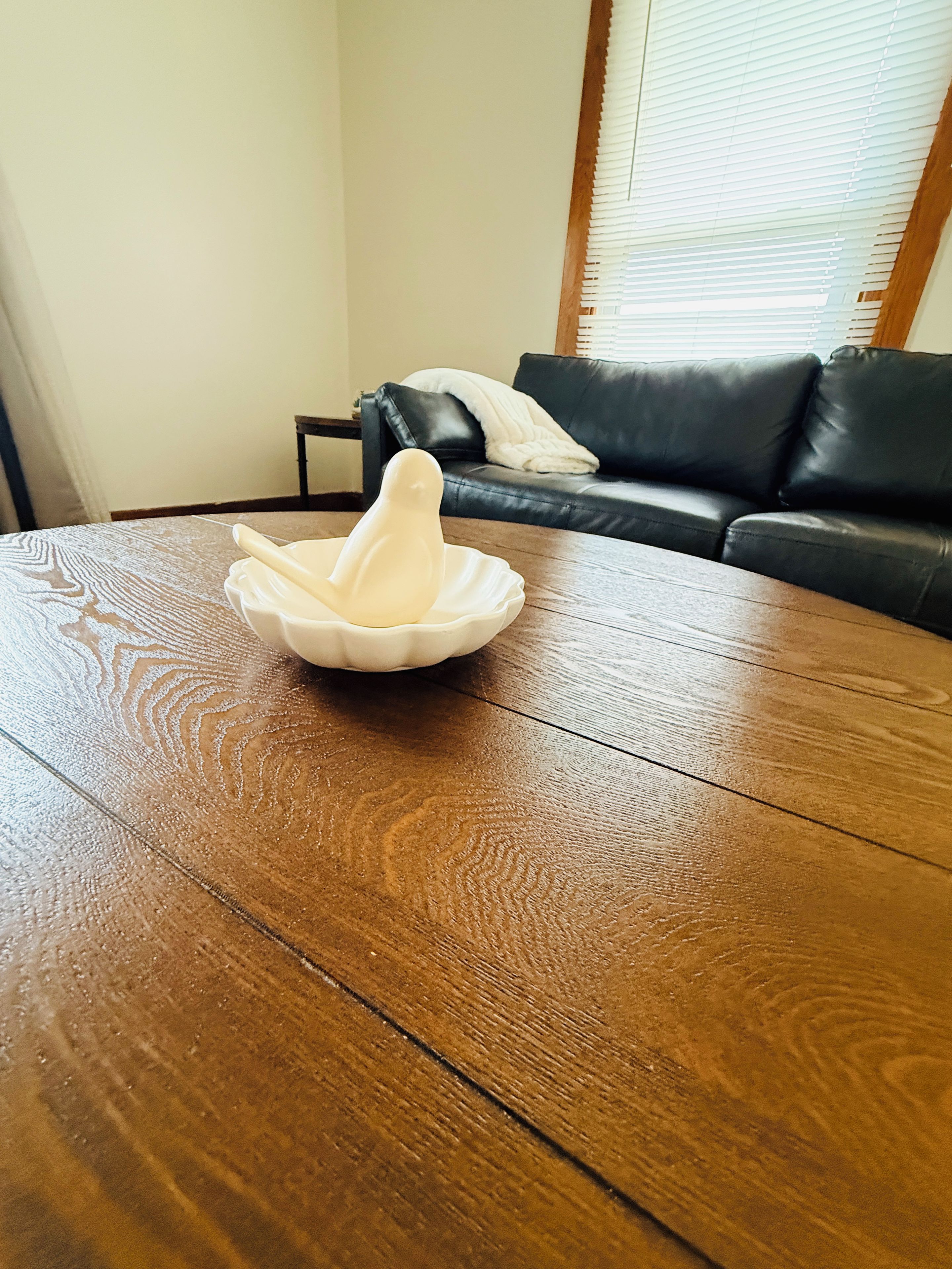 A wooden table with a white object on it, a black leather sofa in the background, and a window with blinds.
