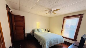 Un dormitorio con cama, mesita de noche, ventilador de techo, ventana con persianas y un mueble para televisor.
