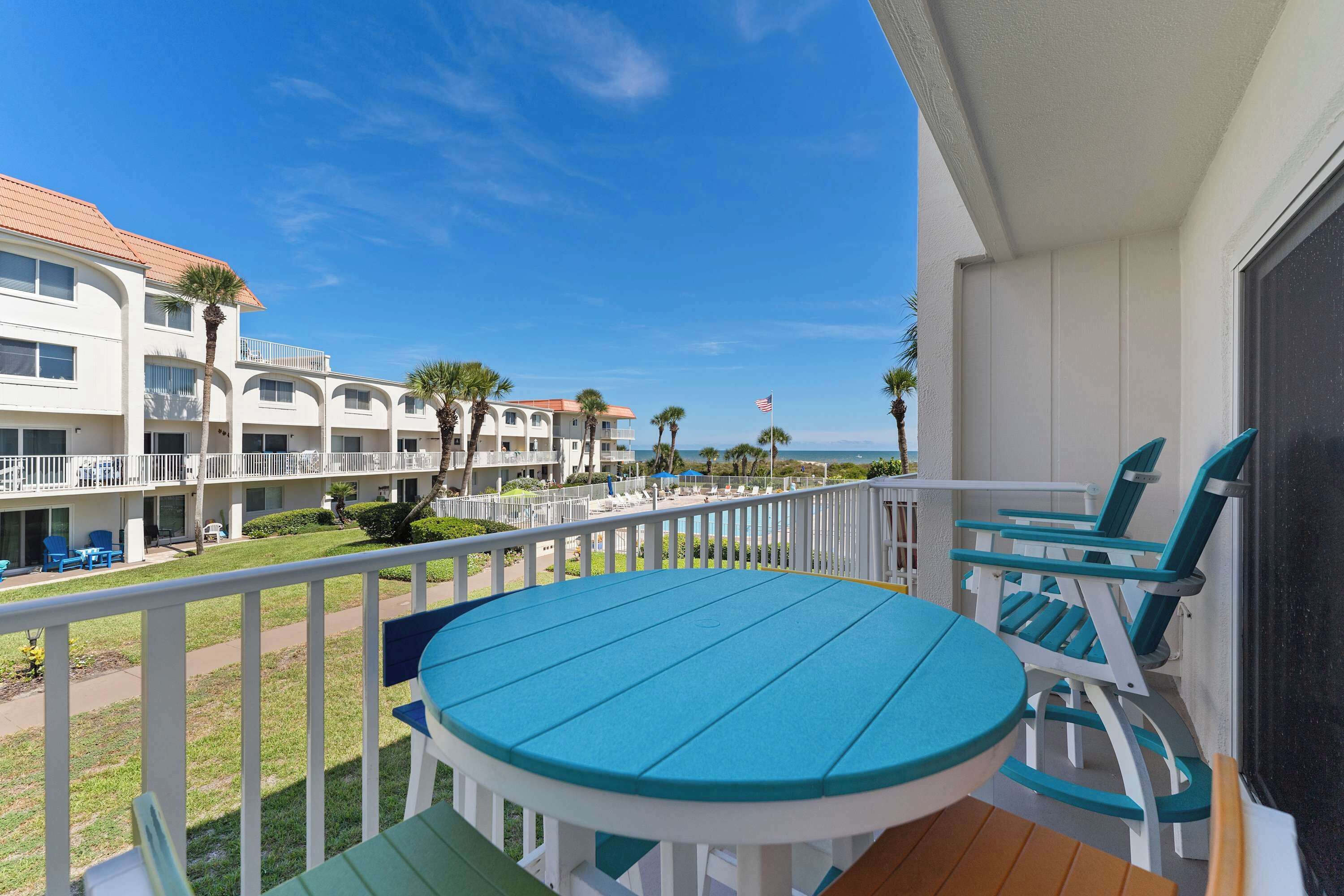 A balcony with a table and chairs, overlooking a pool area and a building with multiple balconies.