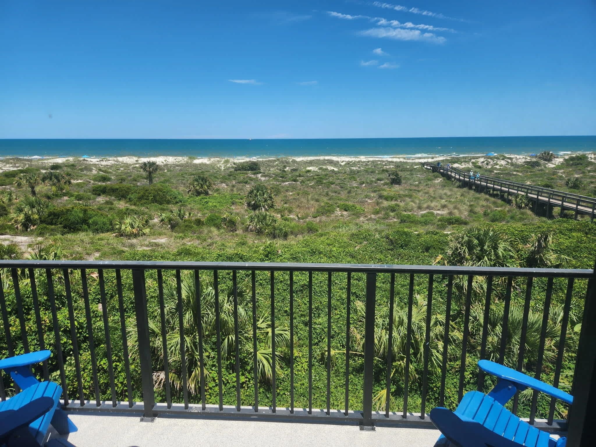 Un balcón con vista a la playa, palmeras y un paseo marítimo de madera.