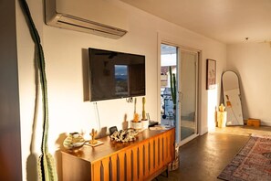A modern living room with a flat-screen TV mounted on the wall, a wooden console table with decorative items, a large mirror, and a sliding glass door leading to an outdoor area. - Friendship House | 5 Acres | Secluded | Hot Tub (Twentynine Palms)