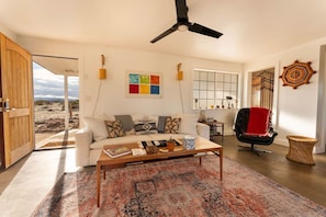 A living room with a sofa, coffee table, and a view of the desert through an open door. - Friendship House | 5 Acres | Secluded | Hot Tub (Twentynine Palms)