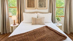 Une chambre à coucher avec un lit, deux tableaux encadrés, une table de chevet avec une lampe et une fenêtre avec des rideaux.