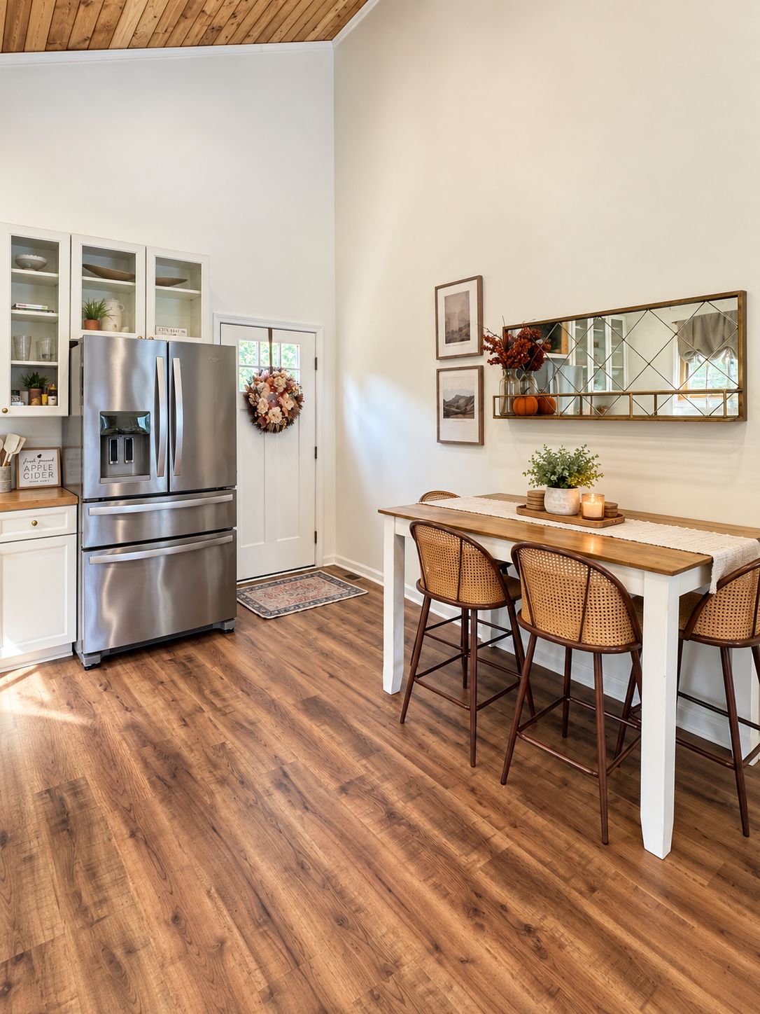 A kitchen with a stainless steel refrigerator, white cabinets, a wooden dining table with four chairs, and a decorative mirror on the wall.