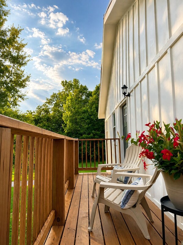 A wooden deck with a white chair and a table, a potted plant with red flowers, and a building with vertical siding. - Private Pool Retreat- Fire Table + Country Escape in Cleveland TN (Cleveland)