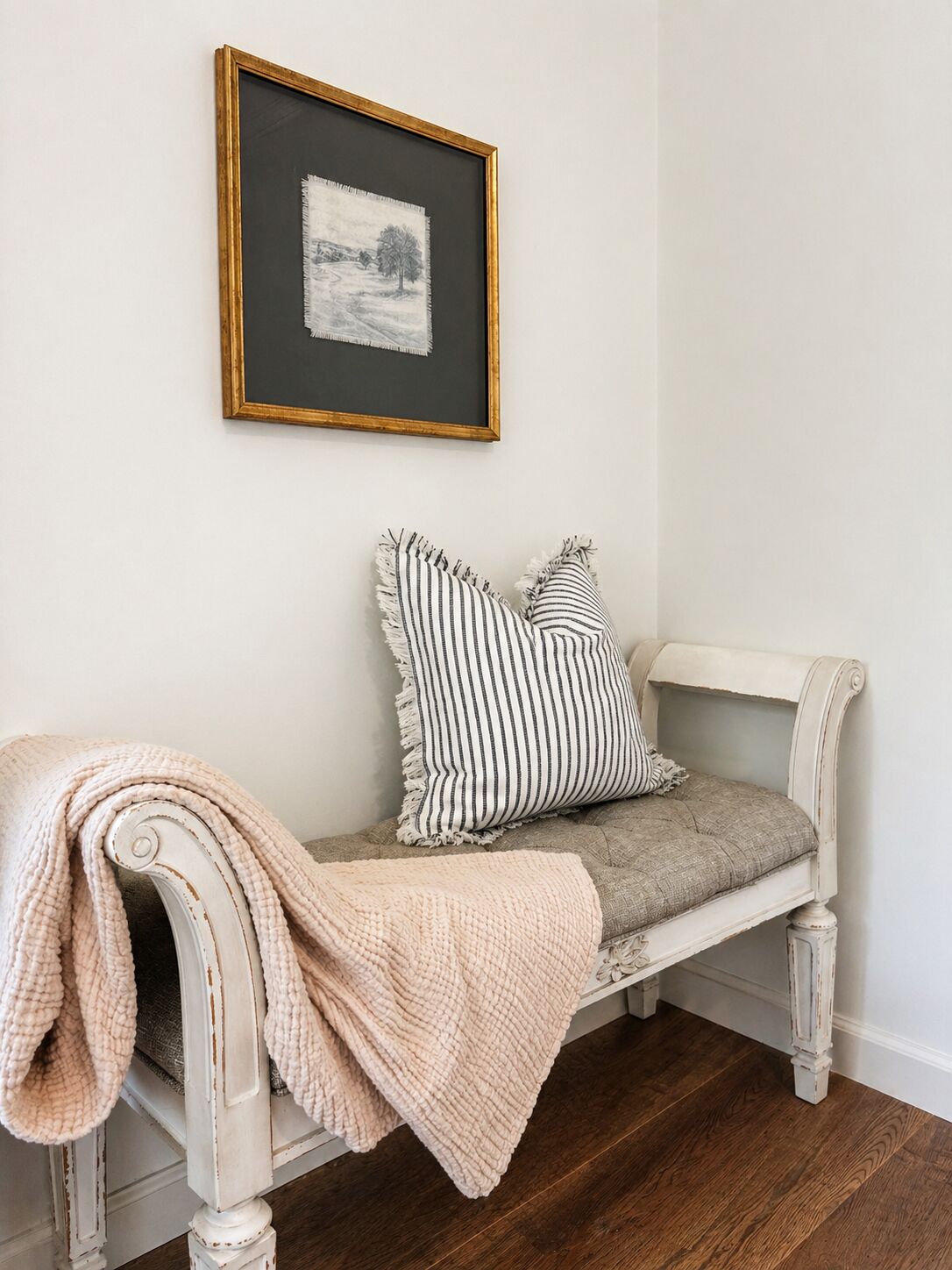 A white bench with a cushion and a throw blanket, a framed picture on the wall, and a wooden floor.