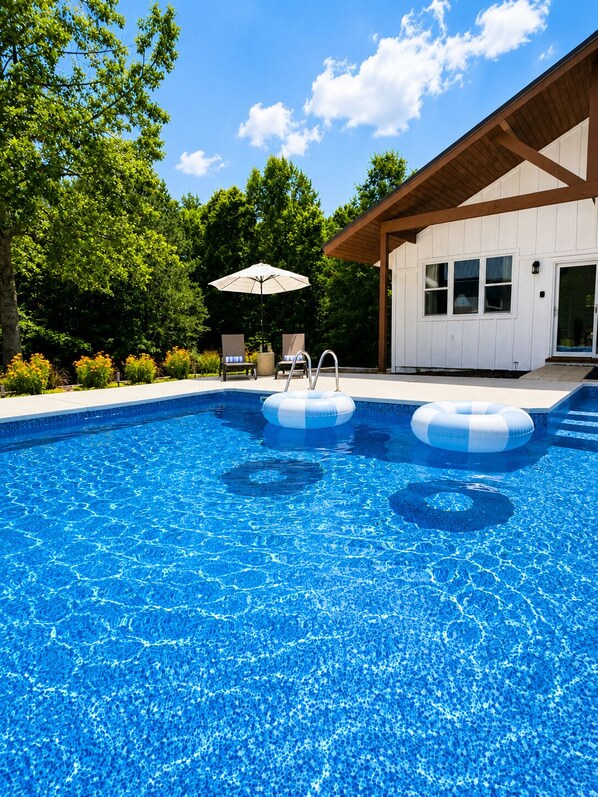A clear blue swimming pool with two inflatable rings, a wooden deck, and a covered seating area with umbrellas.