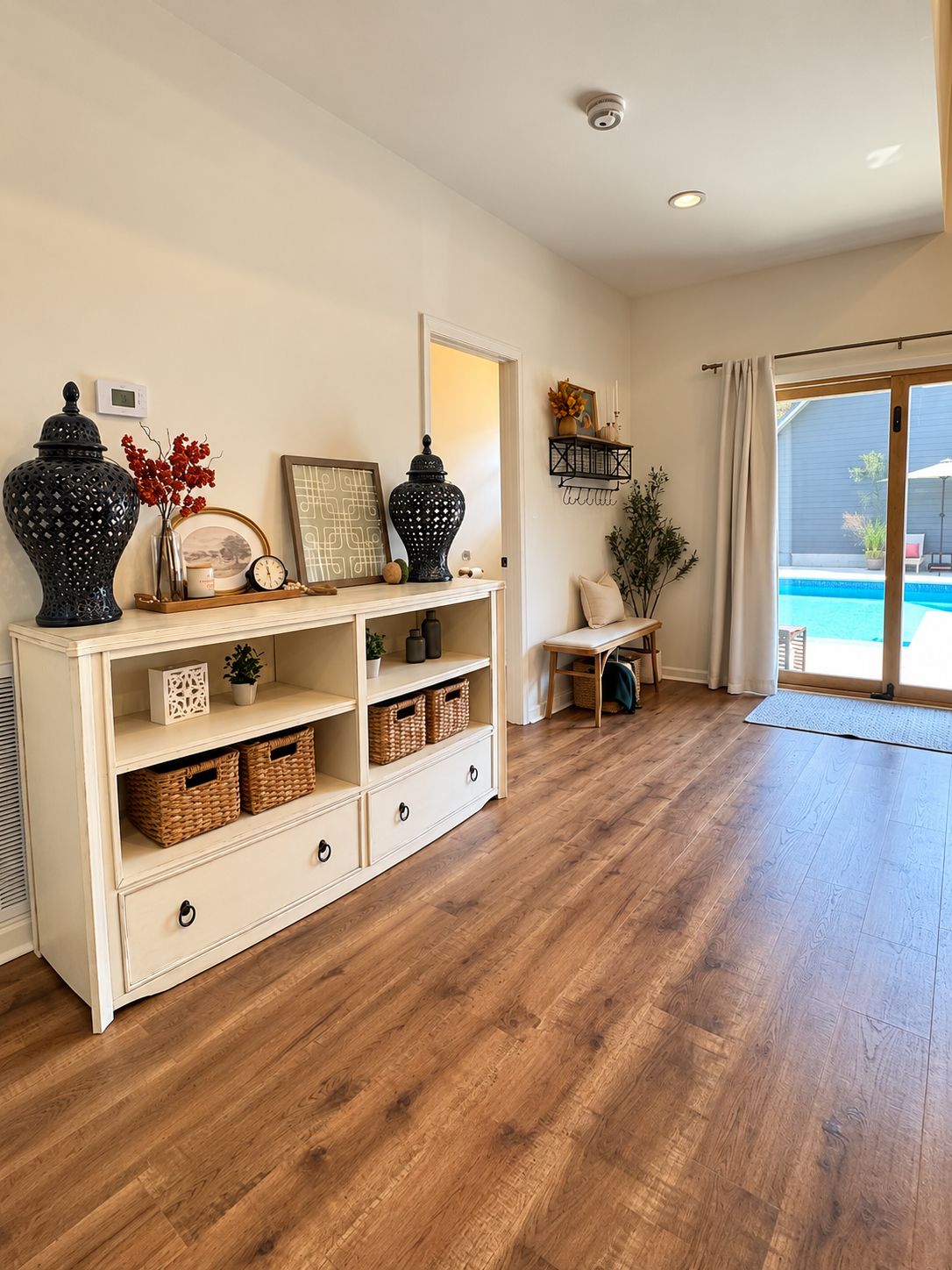 Een kamer met een houten vloer, een witte console tafel met decoratieve objecten, een glazen schuifdeur naar een zwembadgedeelte en een aan het plafond bevestigde beveiligingscamera.