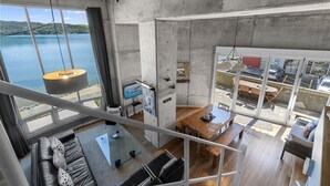 A modern interior with a large window overlooking a body of water, a dining area, and a seating space with a view. - Iconic property • Spectacular outlook (Raglan)
