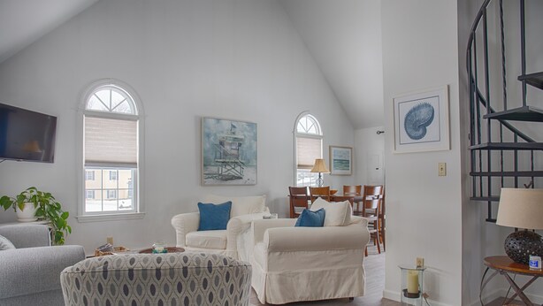 A spacious living room with a high ceiling, white walls, and a staircase. There are two white sofas, a patterned armchair, and a dining area with wooden chairs. - Family-Friendly Wells, Maine Vacation Home Near Ogunquit & Beaches (Wells)