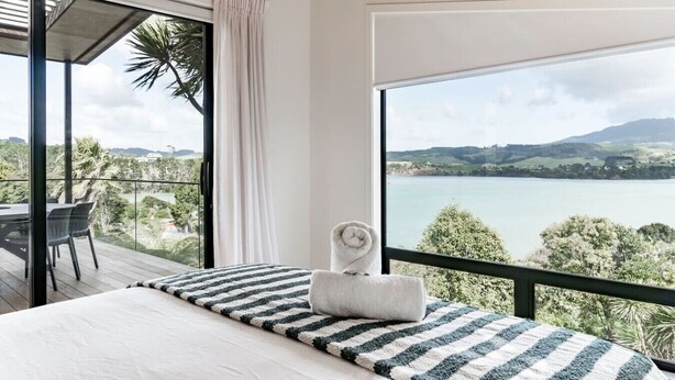 Une chambre avec une grande fenêtre donnant sur un lac et des collines. On y trouve un lit avec un oreiller à rayures et une serviette enroulée posée dessus.