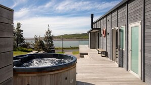 A wooden deck with a hot tub, overlooking a body of water and hills. - Ocean Beach Retreat • Stylish Prime Beachfront (Raglan)