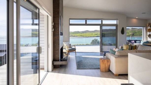 A spacious living room with a large sliding glass door leading to a balcony overlooking a lake and hills. - Ocean Beach Retreat • Stylish Prime Beachfront (Raglan)