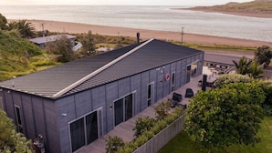 A modern building with a flat roof, large windows, and a patio area with outdoor furniture. - Ocean Beach Retreat • Stylish Prime Beachfront (Raglan)