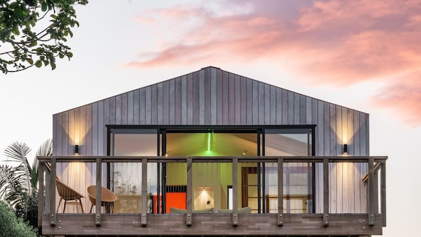 Uma casa moderna de dois andares, com varanda, janelas amplas e interior visível, incluindo um sofá e uma luminária.