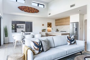 Una sala de estar moderna con un sofá de color gris claro, un comedor con sillas blancas y una cocina con armarios de madera y electrodomésticos de acero inoxidable.