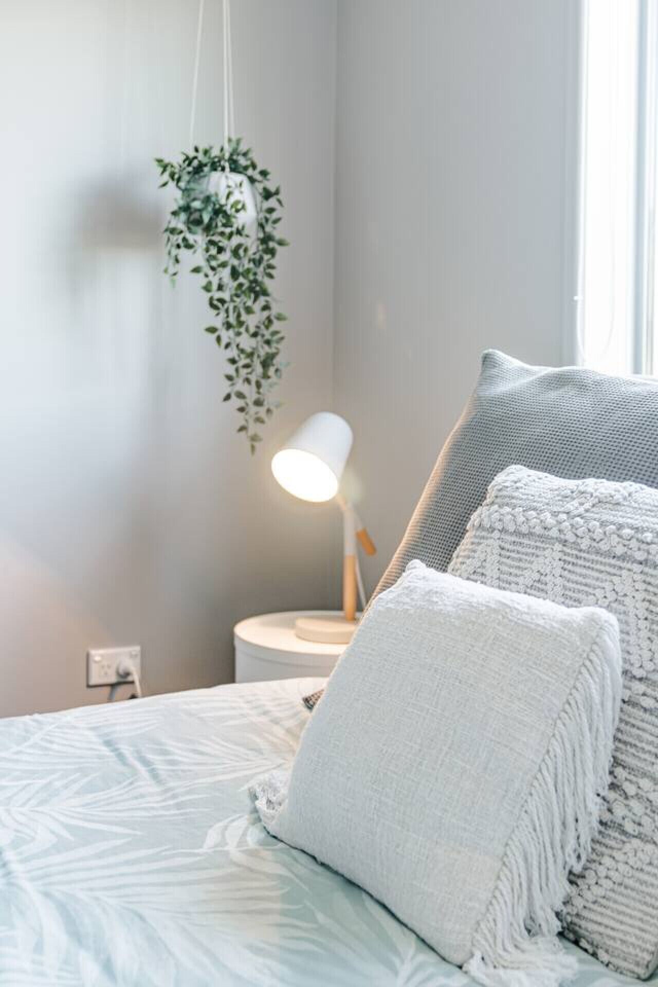 A neatly made bed with white bedding and gray pillows, a bedside lamp, and a hanging plant.