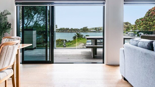 A modern living room with a sliding glass door leading to a deck overlooking a body of water and greenery. - Okiwi Landing • Stylish • Central (Raglan)