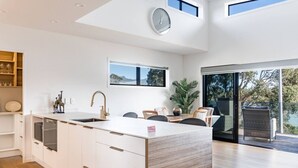 A modern kitchen with a central island, white cabinetry, and a large window offering a view of the outdoors. - Okiwi Landing • Stylish • Central (Raglan)
