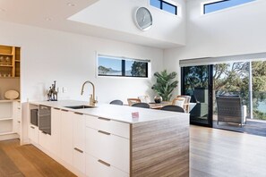 A modern kitchen with a central island, white cabinetry, and a large window offering a view of the outdoors. - Okiwi Landing • Stylish • Central (Raglan)