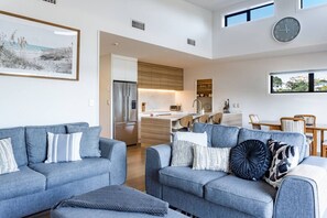 A modern living room with two grey sofas, a coffee table, and a kitchen area with a refrigerator and wooden cabinets. - Okiwi Landing • Stylish • Central (Raglan)