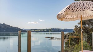 A clear view of a lake with a wooden pier, a beach umbrella, and a lounge area with cushions. - Sunshine Rise • Magical Lorenzen Bay (Raglan)