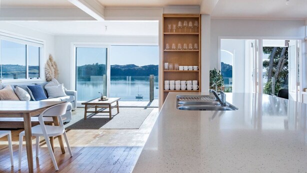 A modern kitchen with a large island, a dining area with a table and chairs, and a view of a lake through large windows. - Sunshine Rise • Magical Lorenzen Bay (Raglan)